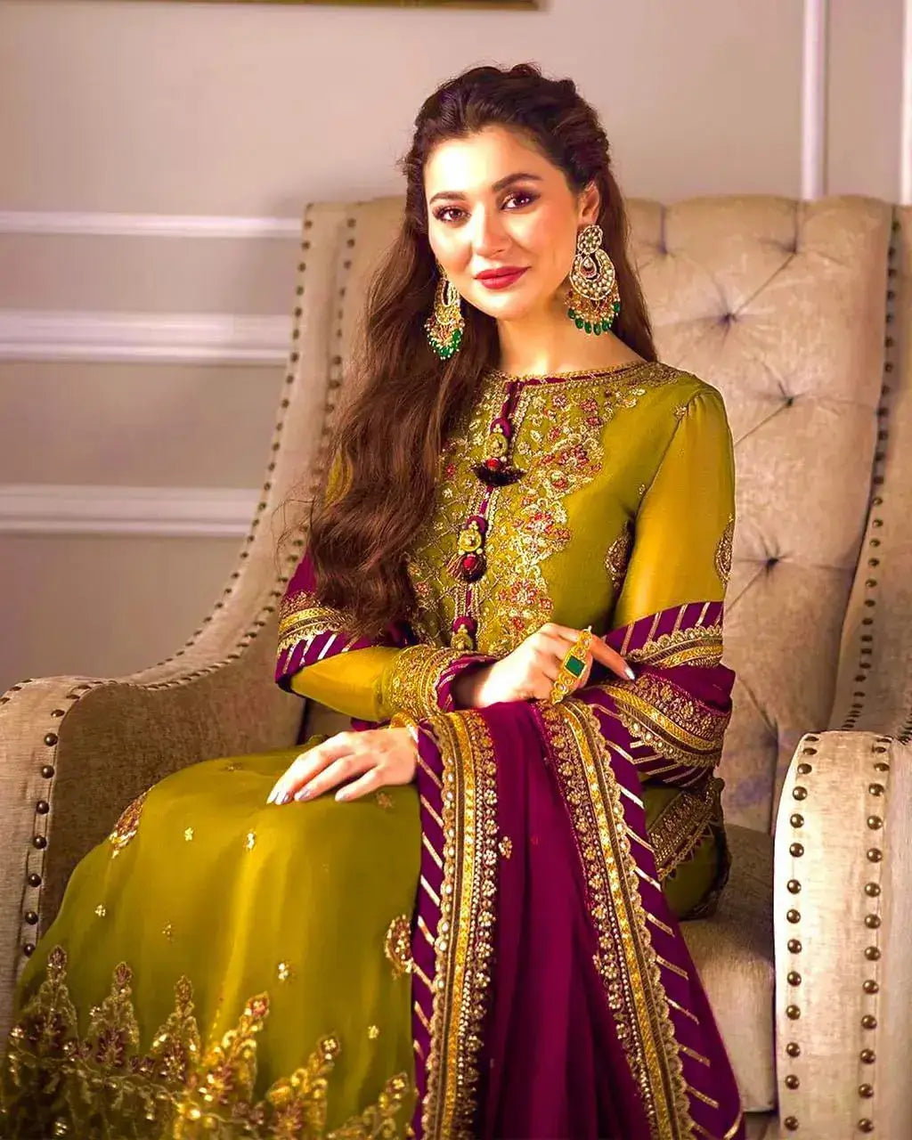 Elegant model showcasing ASIM JOFA 3pc winter wedding outfit in mustard green with burgundy accents, sitting in a stylish chair.